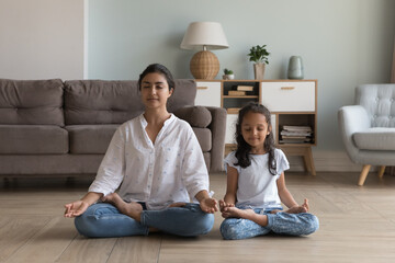 Silent Indian woman and preschooler 5s serene daughter meditating seated in lotus position on warm floor in modern living room. Good life habit, healthy lifestyle, yoga practice with children at home - Powered by Adobe