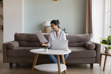 Young happy Indian woman read paper notice with good news smiling seated on sofa at tablet with...