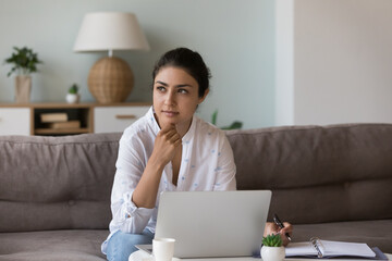 Pensive Indian woman holds pen thinks over ideas staring into distance while sitting on sofa with...