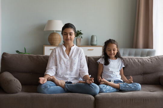 Indian woman and daughter meditating seated in lotus position on sofa at home, closing eyes deep in calm state of mind enjoy yoga practice together in living room. Parent teach child good life habit