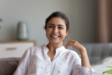 Head shot young Indian woman sit on couch smile look at camera. Happy female rest on comfortable sofa at home laughing enjoy carefree leisure at new modern apartment. Tenant portrait, positive emotion