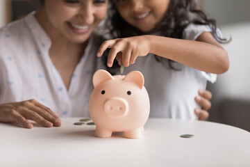 Obraz na płótnie Canvas Smiling Indian mom and her little 6s cute daughter dropping coins into piggy bank. Loving parent teach child be thrifty, to save pocket money for future education or purchases, think about tomorrow