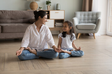 Indian woman laughing seated cross-legged on floor doing meditation practice with cute little daughter at modern cozy home. Loving parent teach child yoga, instill good habits for healthy lifestyle - Powered by Adobe