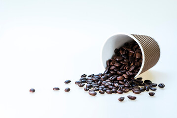 Roasted coffee beans flowing out of a paper cup on a white background.
