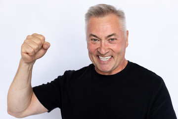 Closeup of joyful senior man with clenched fist. Happy Caucasian male model in black T-shirt with gray hair looking at camera, making winner gesture and celebrating success. Achievement concept.