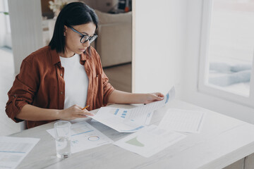 Focused businesswoman works on project, prepares report in charts analyzing documents at home office