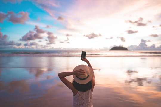 Rear view of young adult tourist asian woman walking relax on beach sand using smartphone for take photo
