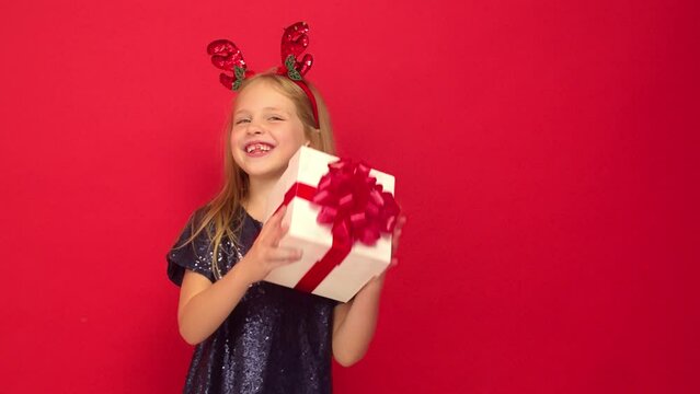Smiling funny child (kid, girl) in Reindeer Hairband. Holding Christmas gift in hand. Christmas concept. Shooting on red background