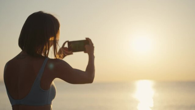 Woman hands holding mobile phone at sunset. Young woman in sport clothes taking photos with her cell phone in a beautiful amazing sunset over sea. Taking a picture on a smartphone during a vacation