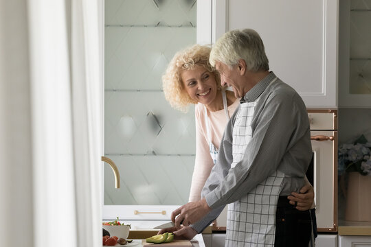 Attractive Older Couple Talking Preparing Vegetarian Dish, Senior Husband Cuts Avocado Enjoy Conversation With Aged Wife Cooking Together Healthy Dish In Modern Kitchen. Routine, Romantic Relationship