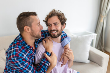 Cheerful young gay couple sitting together. Two affectionate male lovers smiling cheerfully while embracing each other. Young gay coupe being romantic.