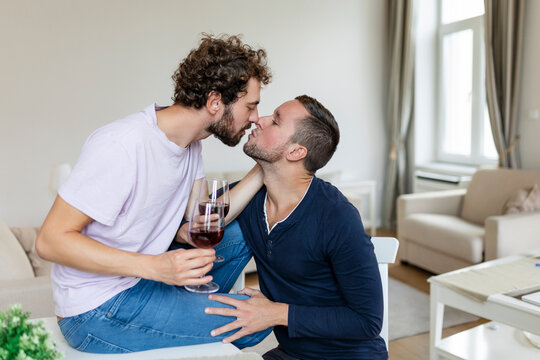 LGBTQ+ couple embracing each other and srinking wine indoors. Two romantic young male lovers looking at each other while sitting together in their living room. Young gay couple being romantic at home.