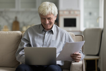 Smiling elderly man sitting on couch put wireless computer on laps use device holds papers make e-payments, pay monthly bills alone at modern home. Older man routine, enough money, finances growth
