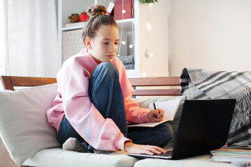 Millennial teen girl sit at couch in living room study on laptop making notes