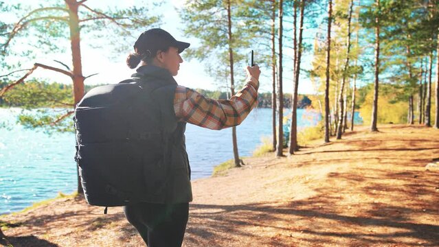 Back view of young woman traveler walking with large backpack on hike in wooded area next to blue lake in outdoors nature landscape. Girl holds phone in hand and using it for photo or navigation.