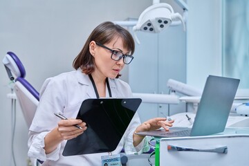 Doctor dentist working in office, using laptop, making notes on clipboard