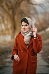 Young brunette girl in orange coat and woolen scarf straightens her headscarf with her hand, looking away, standing against backdrop of autumn park in blur.