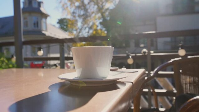 White ceramic cup of hot coffee or tea with steam standing on wooden table of street cafe against sunlight outdoor