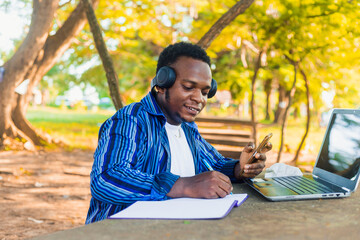 young handsome black male student sitting writing on paper using mobile phone in hand with a...