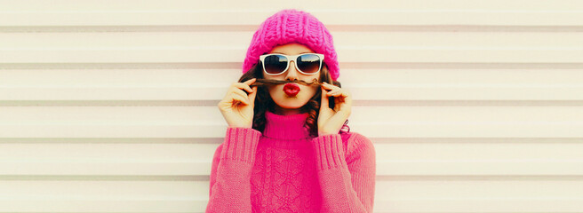 Portrait of funny young woman showing mustache hair blowing her red lips having fun wearing knitted pink hat, sweater on white background - obrazy, fototapety, plakaty