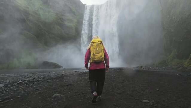 travel adventure hiker traveler walking under skogafoss waterfall,follow steadicam shot of female solo traveller wearing backpack walking to the water stream falling from mountain cliff