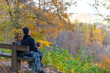 a young woman with a dog on her lap sits on a bench and observes the autumn landscape