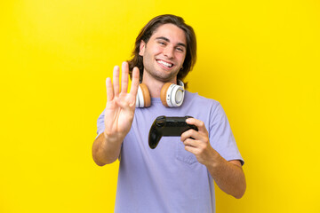 Young handsome caucasian man playing with a video game controller over isolated on yellow background happy and counting four with fingers