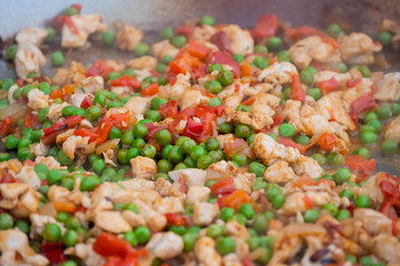 Process of cooking chicken meat pieces, peppers, peas for Spanish paella in huge wok at summer outdoor food market - close up, selective focus. Outdoor cooking, gastronomy and street food concept