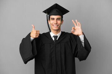 Young Argentinian university graduate isolated on grey background showing ok sign and thumb up gesture