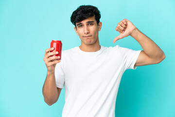 Young Argentinian man holding a refreshment isolated on blue background showing thumb down with...