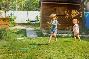 Fun in the garden - boy and girl run around water sprinkler - obrazy, fototapety, plakaty