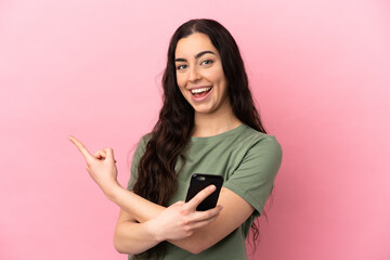 Naklejka premium Young caucasian woman isolated on pink background using mobile phone and pointing back