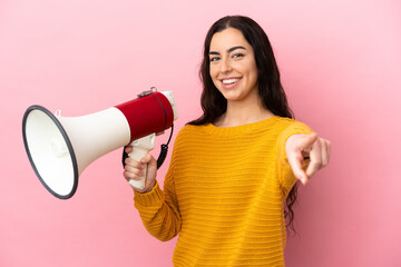 Young caucasian woman isolated on pink background holding a megaphone and smiling while pointing to...