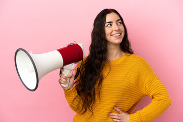 Young caucasian woman isolated on pink background holding a megaphone and looking up while smiling