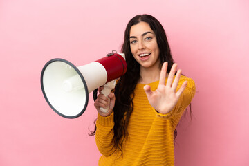 Young caucasian woman isolated on pink background holding a megaphone and saluting with hand with happy expression