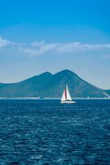Day view of sailing yacht with white sails against a calm sea with calm waves and mountainous landscape background.