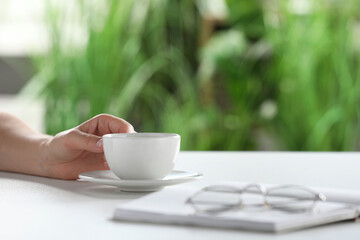 Woman holding cup of coffee at white table indoors, closeup