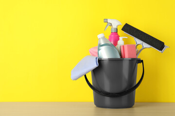 Bucket with different cleaning supplies on wooden floor near yellow wall. Space for text