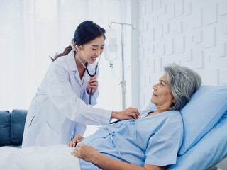A beautiful young Asian woman doctor in white suit smiled while using stethoscope to examine, listen to heartbeat of elderly old female patient in blue dress who was lying on bed in hospital room.