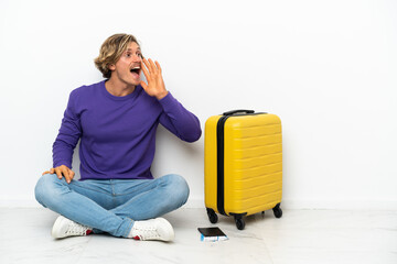 Young blonde man with suitcase sitting on the floor shouting with mouth wide open to the lateral