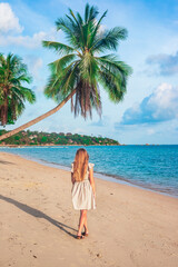 woman with long blond hair in a dress walks along a tropical seashore with palm trees. Travel and tourism