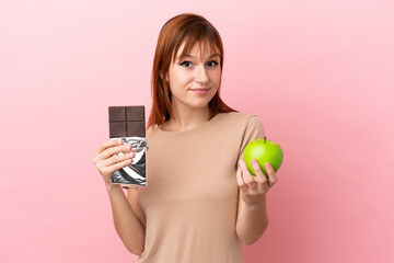 Redhead girl isolated on pink background taking a chocolate tablet in one hand and an apple in the other