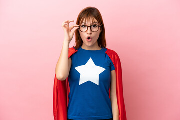 Super Hero redhead girl isolated on pink background with glasses and surprised