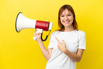 Redhead girl isolated on yellow background holding a megaphone and pointing side