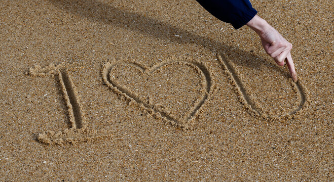 I love you written in the sand of a French beach with a hearth in place of the word love. A white hand finishing to draw the U is visible.
