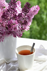 The postcard is beautiful. A mug of tea, an old book and a bouquet of purple lilac. A beautiful still life. Spring time. The concept of Good morning.