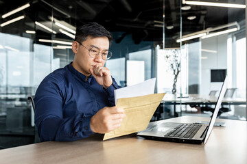 Thinking asian businessman working inside modern office building, man reading letter received from...