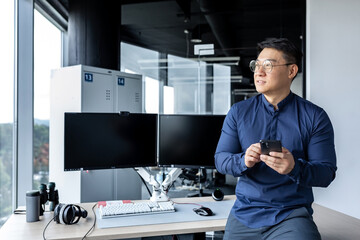 Portrait of serious Asian architect businessman, man looking out window working in modern bright office inside using dual computer, businessman holding phone in hands.