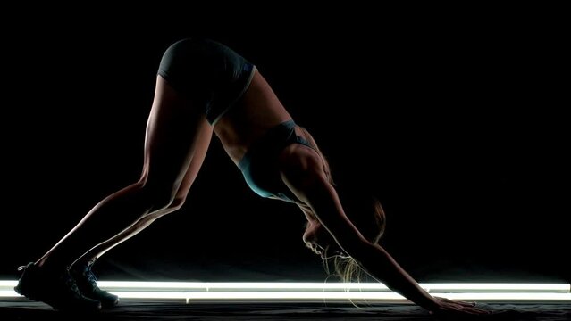 Silhouette female runner stretching and warming up for race. Girl in sportswear posing on lit track against dark background.