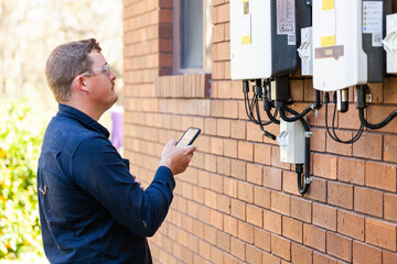 Electrician troubleshooting solar power box on wall of home using phone
